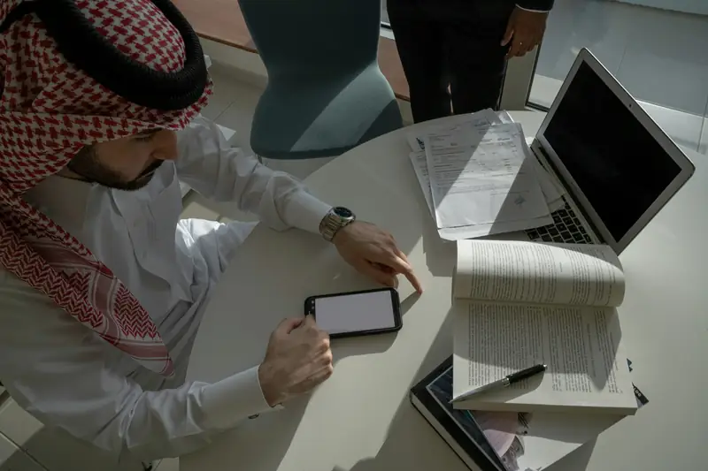 Saudi manager in a shemagh checking a phone beside a laptop